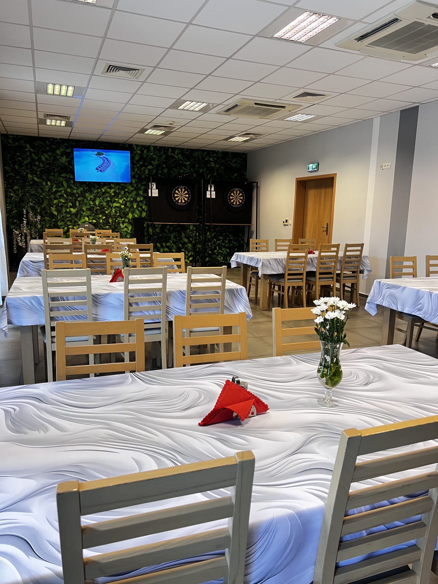 Stoły nakryte do przyjęcia w sali restauracyjnej, w tle ściana z zielenią, telewizor i tarcze do darta